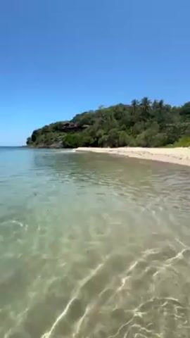 The video shows a tropical beach on a clear, sunny day. The water is shallow, clear, and light blue, with gentle waves lapping against the tan sand. A hill covered in lush green trees and palms rises at the end of the beach. The sky is a clear, bright blue. The camera appears to be at water level.