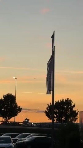 The video begins by showing a flagpole with two flags. The flags appear to be made of a lightweight material, as they are blowing gently in the wind. The sky behind the flagpole is a gradient of colors, with shades of orange, pink, and yellow from the setting sun. There are also some light pink clouds scattered across the sky. In the distance, there is a small airplane visible in the sky, leaving a faint contrail behind it. To the left of the flagpole, there is a tall lamppost with a light on. Further in the background, there are silhouetted trees and the outline of buildings, possibly an airport control tower. In the foreground, the top of several cars parked in a lot are visible. The video is shot from a low angle, looking up at the flagpole and the sky.