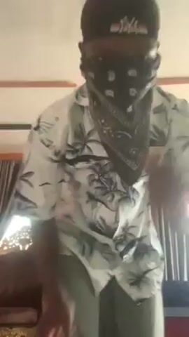 A person wearing a black baseball cap, a gray patterned bandana covering their face, a white patterned short-sleeved button-up shirt, and gray pants is seen in a room with beige walls and white trim. The person moves their arms and head to the music, eventually using both hands to cover their face and pull their hair. A gold-colored ornamental chair is in the background, along with brown curtains. A modern light fixture hangs from the ceiling.