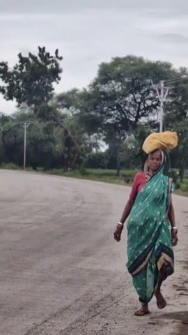 A woman walks along a dirt road carrying a yellow bag on her head. She is wearing a teal and green sari with a blue border and a red shirt. The road is unpaved and appears to be in a rural area with trees and power lines visible in the background. The sky is overcast. A yellow truck drives by in the distance.