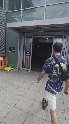 The video shows a person in a purple t-shirt with a skull print and white shorts walking towards an automatic sliding glass door. They are wearing a black fanny pack across their chest. The person has short, dark hair and appears to be entering a building with a modern architectural design, featuring large glass windows and a metal facade. Signs indicating "Departures" with arrows pointing inward are visible on the doors. Inside the building, another person wearing a blue uniform with a yellow scarf and carrying a black suitcase is seen walking towards the camera. Further inside, an individual in an orange jacket and dark skirt is walking away from the camera. The environment appears to be an airport terminal or similar transportation hub.