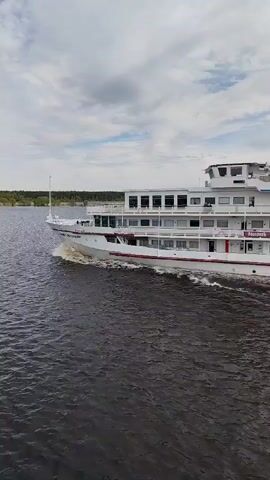 The video shows a white river cruise ship, with "САНКТ-ПЕТЕРБУРГ" (Saint Petersburg) written on its side, moving on a river or lake. The ship has multiple decks and windows. The water appears dark, and the sky is cloudy. The shoreline is visible in the background, lined with trees. The camera appears to be positioned on a bridge or another vessel, capturing the ship as it cruises by.