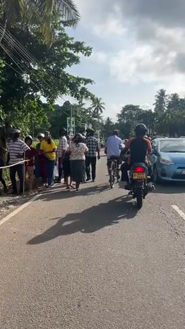 The video shows a street scene in what appears to be a tropical location. A group of people are gathered on the left side of the road. Several individuals are walking along the road.  A person on a bicycle and a person on a motorcycle drive by. There are trees and power lines in the background. The sky is partly cloudy.