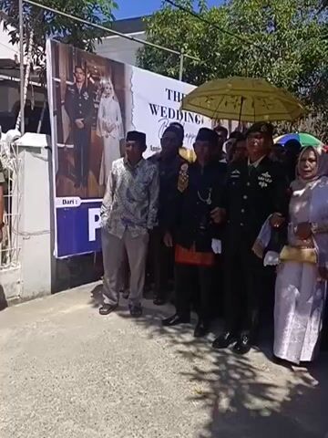 A group of people are gathered outdoors, seemingly for a wedding celebration. In the foreground, a banner is visible, displaying a wedding photograph and text that reads "THE WEDDING" and "Anniversary". Several individuals in formal attire, including a man in a military uniform and a woman in a silver dress and headscarf, are the central focus. Others are dressed in traditional clothing and casual attire. Some are holding umbrellas, suggesting a sunny day. The atmosphere appears festive, with people smiling and interacting. The scene is captured from a slightly low angle.