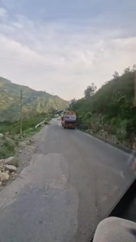 The video shows a truck driving on a winding mountain road. The road is paved and appears to be two lanes wide. The truck is red and yellow and is loaded with what appears to be hay or straw. The road is flanked by steep, green, and brown hillsides. The sky is cloudy and the sun is beginning to set, casting long shadows. There are power lines running along the side of the road. The camera is mounted inside a vehicle, likely a car or bus, and is looking out the front window. The vehicle appears to be moving.