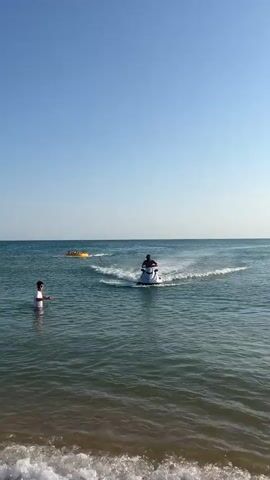 The video shows a sunny day at the beach with clear blue skies. In the foreground, a person in the water is interacting with a jet ski. Further out in the water, a jet ski is being ridden by a person wearing a dark mask and cap. The jet ski is creating a spray of water. Also in the water, a yellow inflatable tube is being pulled by a rope, carrying several people. Another person is standing in the water near the jet ski, holding onto it. The waves are gentle, and the water is a greenish-blue color. The scene suggests a fun recreational activity on the water.