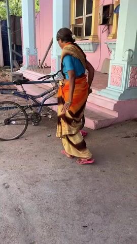 A woman wearing a blue blouse and an orange and brown sari is sweeping with a broom near a bicycle. She is standing in front of a pink and blue structure, possibly a building or a temple, with columns. There is debris on the ground, which she is gathering. A bicycle is parked next to her. The scene appears to be outdoors, with a concrete ground and some vegetation in the background.