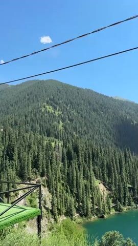 A vertical video shot from an elevated perspective showcases a scenic mountain landscape. The scene is dominated by a steep, densely forested mountainside covered in evergreen trees. A turquoise lake is visible in the lower right portion of the frame, with a road running alongside its edge. In the foreground, there is a railing and a small structure with a green roof. A young woman wearing a baseball cap, sunglasses, and a white t-shirt is captured in several frames, posing and looking towards the camera. The sky is clear and blue, with a single white cloud visible. The video appears to be taken during the daytime.