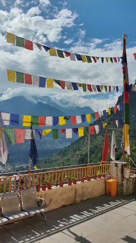The video shows a scenic view from a balcony overlooking a valley and mountains. Prayer flags in various colors are strung across the frame, fluttering in the breeze. In the foreground, there are three empty metal chairs and a low wall with potted plants. The sky is blue with scattered white clouds, and the sun is casting shadows on the ground. The overall impression is one of peace and tranquility, with a sense of spiritual significance due to the prayer flags.