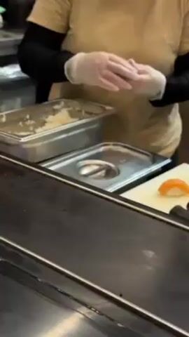 A person wearing a tan shirt and black sleeves, and donning transparent gloves, is preparing food behind a counter. A metal tray filled with white rice is on the left. Next to it is a smaller metal tray with a circular indentation. A white cutting board holds a piece of salmon nigiri and two rolls of sushi wrapped in black nori. The person picks up the salmon nigiri. The person prepares more food while standing at the counter.