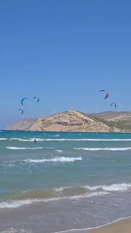 A wide shot of a beach on a sunny day. The ocean is a vibrant turquoise with white-capped waves rolling onto a sandy shore. In the distance, a hilly, rocky coastline is dotted with wind turbines. Several kitesurfers are visible in the water, their colorful kites dotting the blue sky. A few people are on the beach, some swimming or wading in the water, others relaxing on the sand. The sky is a clear, cloudless blue.