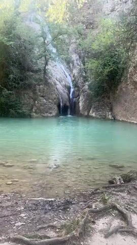 The video shows a waterfall cascading into a pool of turquoise water. The waterfall is surrounded by lush green foliage and rocky cliffs. The water in the pool is clear, and the bottom is visible in some areas. The sunlight filters through the trees, creating a serene and beautiful atmosphere.