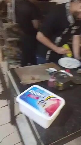 A man with a beard, wearing a black shirt with a decorative collar, is in a kitchen. He is holding a yellow squeeze bottle and appears to be squeezing something onto a white plate. The plate has some writing or drawings on it. In the foreground, there is a white tub of ice cream labeled "Fanice." To the left of the plate is a wooden cutting board. To the right of the plate, there is a pizza cutter and a small red bottle. The background shows kitchen equipment and utensils. The man is bent over the plate, focusing on his task.