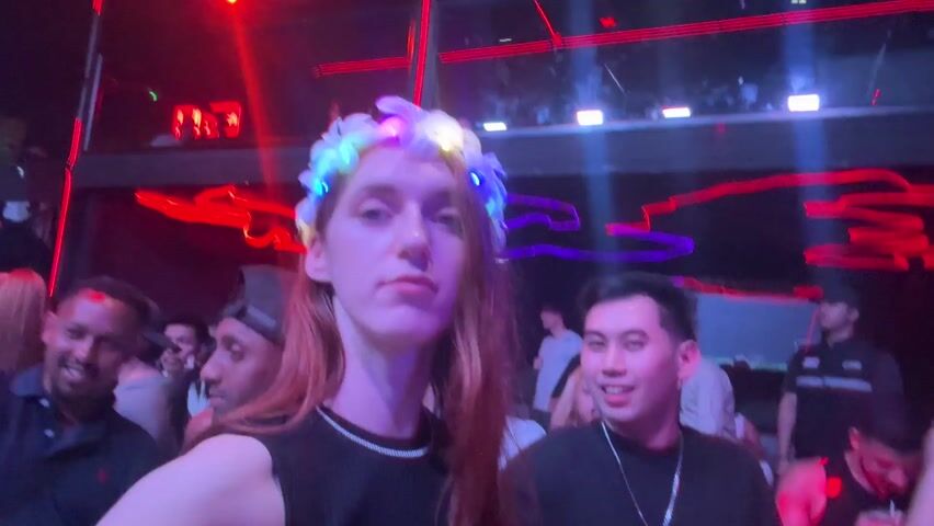 The video shows a crowded nightclub with people dancing and socializing.  A person wearing a glowing flower crown is filming themselves in the foreground, while others dance and move around them. The club has colorful neon lights and a sign that says "CLUB". The atmosphere is dark and energetic.
