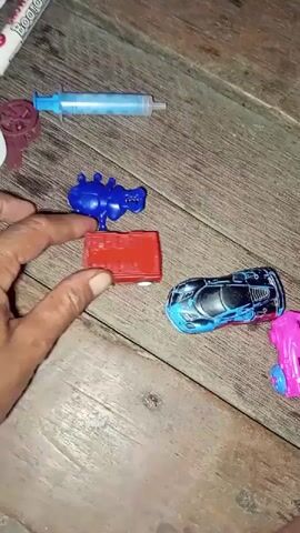 The video shows a person's hands arranging small toys on a wooden surface. The toys include a blue syringe, a red rectangular block, a blue insect-like figure, a blue and black toy car, and a pink toy car. The person's hands are moving the toys, stacking them, and placing them next to each other. The video captures a close-up view of the toys and hands.