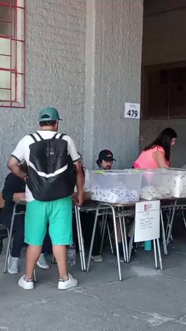 The video shows a polling station. A man in a green cap and shorts stands by a table, and then an older man in a patterned shirt approaches the table. Three election workers sit behind the table, sorting through ballots. There are plastic bins on the table filled with ballots. A sign on the wall says "Mesa 479". The scene takes place in an outdoor area with gray walls.
