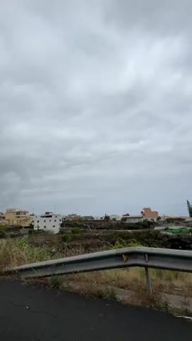 A low-angle video shows a cloudy sky and a rural landscape with houses on a hillside. A metal guardrail is visible in the foreground, with dry grass and vegetation growing alongside it. A traffic sign with a red circle is visible on the left side of the frame. Telephone poles and wires are also present. The houses are predominantly white and yellow and are built on a terraced hillside. The overall scene suggests a moderately overcast day in a somewhat underdeveloped area.