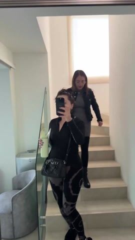Two women are walking down a set of bright, white stairs. The woman in the foreground is taking a selfie with her phone. She is wearing a black long-sleeved athletic top and tie-dye leggings, and has a black purse slung over her shoulder. She's also holding a light green water bottle. The woman behind her is wearing a grey dress, black denim jacket, and black boots. She is also holding a phone. They are walking down the stairs indoors. There is a mirror to the left, reflecting a grey chair and what appears to be a water dispenser. Natural light streams in from a window at the top of the stairs.