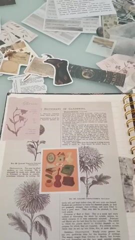 A high-angle shot shows a spiral-bound scrapbook open on a cluttered surface. The surface is covered with stickers, printed quotes, and paper cutouts. The scrapbook pages are decorated with vintage-style botanical illustrations, stickers, and handwritten text on a page featuring a purple galaxy design. The overall impression is one of a creative workspace dedicated to journaling or scrapbooking.