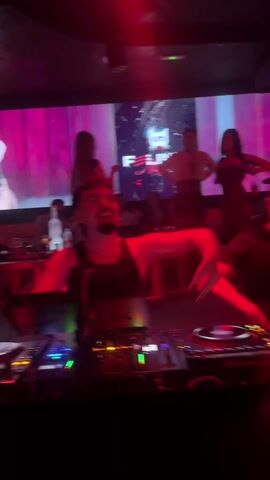 Here's a description of the video:

The video shows a DJ performing at a nightclub. The DJ is a man with dark hair and a goatee, wearing a black tank top. He is standing behind a Pioneer DJ console. Red lights fill the space. Behind him is a video screen displaying various images, including a woman in a mask and the club's logo. Several people are standing and dancing behind the DJ. One man wears a white suit and holds a large horn-shaped instrument. There are tables with drinks in the background, and the overall atmosphere is that of a vibrant nightclub scene. The camera angle is from the DJ booth, giving a first-person view of the performance.