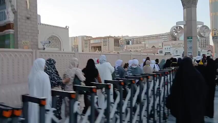 A crowd of people, mainly women wearing hijabs and abayas, are walking along a pathway lined with metal barriers. Some of them are taking pictures or videos with their phones. In the background, there's a building with arched windows and a minaret, typical of Islamic architecture. There are also modern buildings and structures with decorative elements like wind catchers. The sky is a pale blue, suggesting it's either early morning or late afternoon. The scene appears to be taking place near a religious site, possibly in Saudi Arabia.