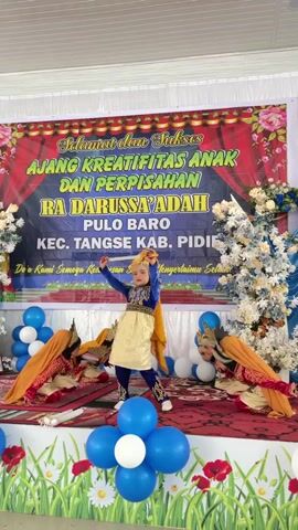 A group of young girls in traditional attire are performing on a stage. A banner behind them reads "Selamat dan Sukses" and "AJANG KREATIFITAS ANAK DAN PERPISAHAN RA DARUSSA'ADAH PULO BARO KEC. TANGSE KAB. PIDIE." The girl in the center, dressed in a blue and gold outfit, holds a cylindrical object and appears to be the main performer. The other girls are seated around her, also in traditional clothing with orange headscarves. The stage is decorated with blue and white balloons and floral arrangements. The background is a plain wall. The lighting suggests an indoor event.