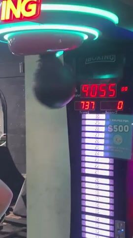 A young person wearing red boxing gloves stands in front of a "BOXING" arcade game. The game has a digital display showing "High Score 9055". The person is in the process of punching the hanging black ball. The game's lights are bright, cycling through green, red, and blue. The background appears to be an indoor gym or arcade, with some gym equipment visible to the left.