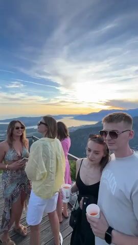 The video shows a group of four people standing on a wooden platform overlooking a bay with mountains in the background. The sky is blue with some clouds and the sun is setting, casting a warm glow over the scene. A young couple in the foreground is holding drinks. The man is wearing a light-colored t-shirt and sunglasses, while the woman is wearing a black top. To their left, two other women are visible. One is wearing a yellow shirt and white shorts, and the other is wearing a patterned dress. The woman on the far right, facing away from the camera, is wearing a red crocheted top and sunglasses and is smiling. The bay below is dotted with boats, and a town is visible along the shoreline. The overall atmosphere is relaxed and celebratory.