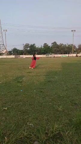 A person in a red dress and black hijab runs across a grassy field. The field appears to be a sports field with lights and a fence in the background. The sky is overcast.