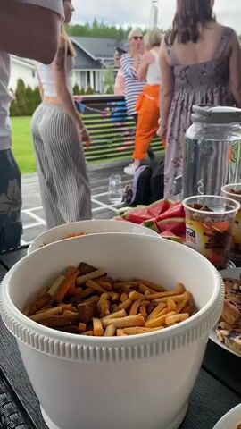 This media file captures a casual outdoor gathering, likely a backyard party, on a cloudy day. In the foreground, a large white bowl is filled with assorted croutons or breadsticks, suggesting food is a central element of the event. Beside it, sliced watermelon and another container of snacks are visible, hinting at a relaxed, communal meal.

The background features a group of people on a deck. One person, seen from the back, wears a white crop top and vertically striped wide-leg pants, with a striped bandana on their head. To their right, another person in bright orange pants and a white tank top is engaged in conversation. Further back, two more individuals are visible, one in a striped top and the other in a floral print dress. The overall atmosphere appears to be social and informal.

Behind the deck, houses with dark roofs are visible amidst lush green trees, indicating a suburban or semi-rural setting. The sky is overcast, suggesting it might be late afternoon or early evening, but there are no strong indicators of specific time. The general impression is one of a pleasant, unpretentious social occasion taking place in a residential area in Joensuu, Finland.

The only visible text is on a small orange and brown package in the background, partially obscured, which reads "Bread Sticks".
