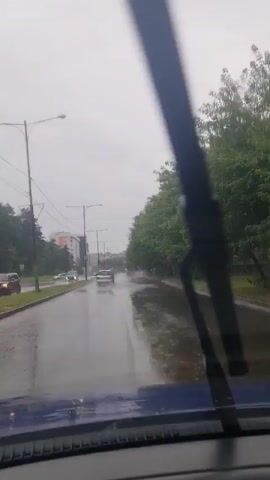 The video shows a view from inside a car driving on a wet road. The road is lined with trees on the right side and buildings and trees on the left side. There are puddles on the road and it appears to be raining lightly, as indicated by the windshield wipers. Several cars are visible on the road, driving in the same direction as the camera. The sky is overcast and gray.
