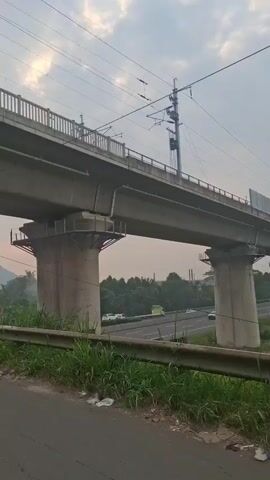 Here is a description of the video:

The video shows a concrete overpass with elevated tracks. Below the overpass, a highway with cars driving on it is visible. The camera moves from underneath the overpass and then onto a narrower bridge with metal railings. A white van is driving over the bridge in the distance. In the background, hills and a telecommunications tower can be seen.
