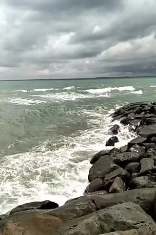 The scene shows a rocky shoreline on a cloudy day. The water is choppy, with waves crashing against the rocks. The sky is overcast with gray clouds. The water is a blue-green color, and a thin line of land can be seen in the distance. There are no people visible in the scene.