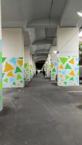 The video shows a covered walkway under an elevated structure, with a gray asphalt surface and white support columns decorated with colorful geometric shapes. Several people walk in the distance, and a person on a bicycle rides toward the end of the walkway. A parked car is visible to the right, and the lighting is somewhat dim.