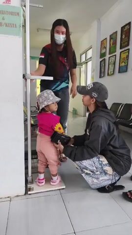 A young child stands on a scale, facing forward and wearing a pink shirt, light-colored pants, a patterned hat, and pink shoes. Another person, wearing a dark hooded jacket, patterned pants, a baseball cap, and black shoes, kneels beside the child, holding a black object. A third person, wearing a black t-shirt with a colorful design, gray pants, and sandals, stands behind the child, adjusting a metal measuring device. The scene takes place indoors, possibly in a clinic, with a wall displaying several colorful framed pictures and a sign that reads "LABORATORIUM & KUSTA". The floor is tiled.
