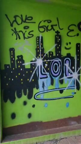 The video shows a vibrant green wall covered in colorful graffiti. The graffiti includes a cityscape, the word "LOL" in large letters with a dripping effect, and various cartoon-like characters, including a ladybug and a green blob. There is also text written on the wall, such as "love this girl" and "hey. jahah." Part of the wall appears to be a segment of a train car with red-framed windows. A light fixture is attached to the wall. The bottom of the wall is bordered by a brown and green strip, and a brick floor is visible in the foreground. The camera pans across the wall, showcasing the different graffiti elements. There are no people visible in the video.