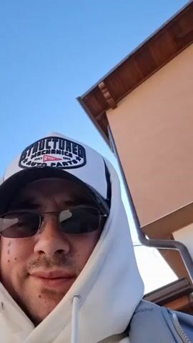 A man in sunglasses and a white hoodie with the hood up is facing the camera. He's wearing a white baseball cap with a black bill and a "Structured Mechanics Auto Parts" logo. He has facial hair and a small mark on his left cheek. The background includes a tan building and a clear blue sky with some power lines. The camera angle is slightly low, looking up at the man. The video is well-lit and the man is smiling slightly.