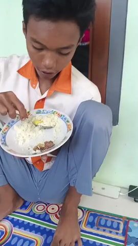 The video shows a young person wearing a white and orange school uniform sitting on the floor and eating rice with a spoon. They are holding a white plate with rice and some other food items. The person is looking at the camera and chewing. In the background, another young person is visible sitting and eating. The environment appears to be outdoors, with a wall and a window visible. There is also a motorcycle in the background. The video is short, and the action is simple, focusing on the act of eating.