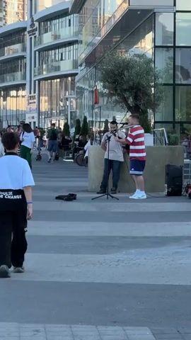 A man plays an acoustic guitar while another man sings into a microphone, both standing on a paved area. The guitarist wears a red and white striped shirt and blue shorts, while the singer wears a tan t-shirt and jeans. They are positioned in front of a large building with glass windows and an olive tree. Other people are visible in the background, some seated at tables and others walking. A black speaker is placed to the right of the performers. The overall environment appears to be an urban outdoor setting, possibly a plaza or promenade, during daylight.