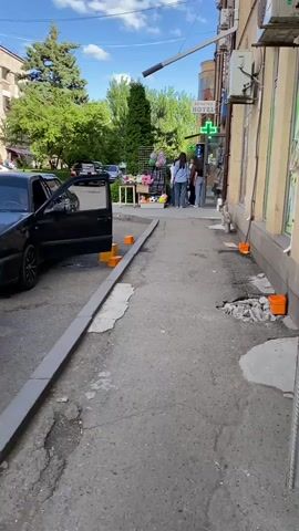 The video shows an outdoor scene on a sidewalk in front of a building. A black car with its driver's door open is parked on the street next to the sidewalk. There are orange traffic cones placed along the edge of the sidewalk. In the background, several people are standing near a small stand displaying various items. A pharmacy with a green cross sign is visible on the building. The sidewalk appears to be in disrepair, with cracked pavement and some debris. Trees and greenery are present in the background, suggesting a daytime setting with sunlight.