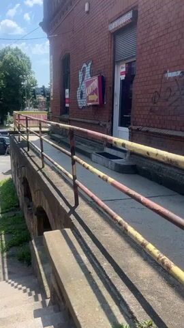 The video shows a street scene in front of a brick building. There is a ramp with a metal railing leading up to the building. A car is parked on the street, and two people are walking in the background. The building has a sign that says "KANTOR" with an arrow pointing to the right. The sky is blue with some clouds.