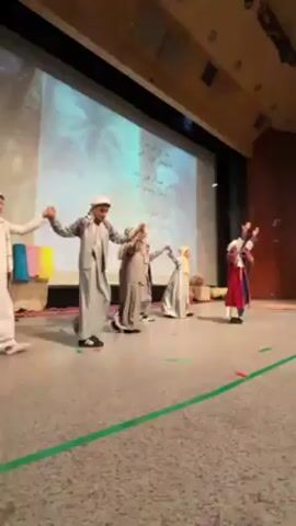 A group of young people is performing on stage in front of a large screen. The screen displays images in Arabic script. Most of the performers are dressed in robes and headwear, while one is wearing a blue and red robe. They walk and dance, holding hands in a line, and raise their arms. The stage has green lines on the floor, and there are some objects on the left side. The hall appears to be a theater with wooden details.