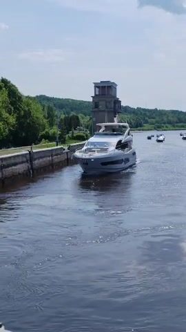 The video shows a gray and white yacht cruising through a narrow waterway, guided by the concrete banks of what appears to be a canal or lock. The sky is a clear blue with scattered white clouds, suggesting a bright, sunny day. The waterway is surrounded by lush green trees and a low, rolling landscape. In the background, there is a tall, three-tiered concrete tower-like structure, possibly a control tower for the canal or a navigational aid. Other boats are visible in the distance, further along the waterway, suggesting there is active boat traffic.