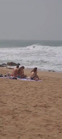 Three women in bikinis are sitting on a towel on a sandy beach. They appear to be looking at their phones. The ocean is behind them, with waves crashing near some rocks. The sky is overcast.