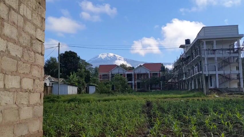 The video shows a view of Mount Kilimanjaro in the background, with buildings under construction in the foreground. The mountain is snow-capped and partially obscured by clouds. There is a field of crops in front of the buildings. The sky is blue with scattered clouds. A section of an unfinished brick building is visible on the left side of the frame. Power lines stretch across the sky.  Small sheds or outbuildings are visible among the trees.