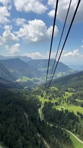 This is a first-person perspective video taken from a cable car descending a mountain in the Dolomites, near Ortisei, Italy. The cable car is moving down towards a valley.

The scene is a breathtaking mountain landscape. In the foreground, a dense forest of dark green coniferous trees covers the steep slopes. Below the forest, a lush green valley unfolds, dotted with scattered buildings, some appearing to be farmhouses or chalets, and crisscrossed by winding roads and paths. In the distance, layers of majestic, rugged mountains rise, their peaks partly obscured by a bright blue sky filled with fluffy white clouds. The sun is shining, indicating it is likely daytime, probably late morning or early afternoon given the brightness and the angle of the light. The weather appears clear and pleasant, ideal for outdoor activities.

The activity taking place is a scenic cable car ride, offering panoramic views of the alpine scenery. The cable car itself is not fully visible, but its cables are a prominent feature in the frame, stretching diagonally across the sky. A small, white cable car cabin can be seen in the distance, suspended from the cables and moving down the mountain.

The overall mood is one of awe and tranquility, capturing the beauty and grandeur of the natural environment. There are no people clearly visible in the frame, nor any discernable text. The focus is entirely on the spectacular natural vista and the experience of traveling through it via cable car.