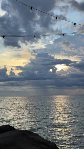 This media file shows a scenic view of the ocean at sunset. The sun is peeking through dramatic, dark clouds, casting a golden light on the water. The sky is a mix of blues, grays, and yellows, with some light rays visible descending from the clouds towards the sea. In the foreground, a dark, rocky shoreline is visible. String lights with exposed bulbs hang across the top of the frame, suggesting an outdoor dining or gathering area. The scene evokes a sense of tranquility and natural beauty. The time of day is clearly sunset, with the weather appearing to be partly cloudy or experiencing a brief shower. The location is identified as Ko Tao Subdistrict, Thailand. There are no visible people or text in the media file.