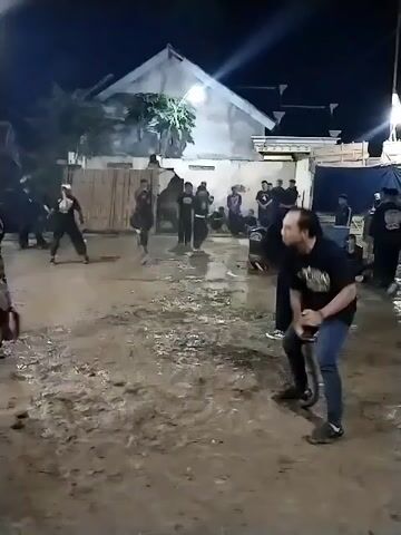 The video depicts an nighttime outdoor performance or ritual in Jenggrik, Indonesia. The setting is an unpaved, muddy ground area, illuminated by bright artificial lights. In the background, a building with a white facade and a prominent triangular roof is visible, along with temporary fencing or scaffolding.

Multiple people are present, mostly men. In the foreground, a man in a black T-shirt, blue jeans, and sneakers is seen holding and maneuvering a whip. To his left, another man, also in dark clothing and sneakers, dramatically falls to the muddy ground. He then begins to spin rapidly on his hands and feet, moving in a circular motion with great agility. Other spectators and participants are visible around the perimeter, watching the activity. The performance is accompanied by traditional Indonesian music, featuring percussion instruments that give it a ritualistic feel. The man spinning appears deeply engrossed in the performance, possibly in a trance-like state, as he executes his fast, continuous rotations.