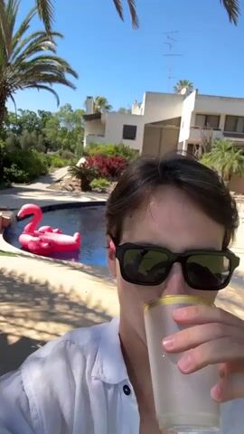 A young person wearing sunglasses and a white shirt takes a selfie while holding a drink. The person is standing outdoors in a sunny environment. Behind them, a swimming pool with a pink flamingo float is visible, along with palm trees, lush greenery, and a multi-story building. The person sips from their drink and smiles at the camera.
