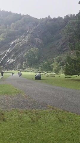 The video pans to the right showing a group of people walking on a gravel path in front of a mountain with a waterfall cascading down. A golf cart is parked on the right side of the path. The scene is green and lush with trees on the mountain and in the foreground. The weather appears overcast.